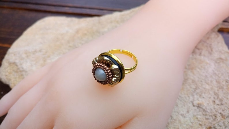 May include: A gold-toned adjustable ring with a floral design. The ring features a central gray stone surrounded by a copper-colored metal, a black band, and a gold-colored adjustable band. The ring is displayed on a mannequin hand.