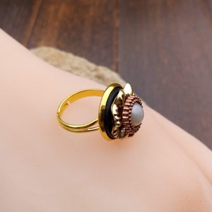 May include: A gold-toned adjustable ring with a decorative setting. The setting features a central gray stone surrounded by layers of copper-colored metal, black accents, and a gold-colored frame. The ring is displayed on a light skin tone.