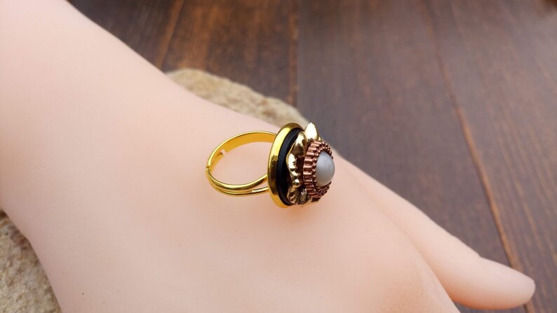 May include: A gold-toned adjustable ring with a decorative setting. The setting features a central gray stone surrounded by layers of copper-colored metal, black accents, and a gold-colored frame. The ring is displayed on a light skin tone.