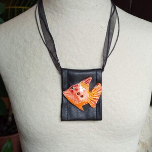 Pu&ograve; includere: Una collana in pelle nera con un piccolo sacchetto nero con un design di pesce arancione e rosso.