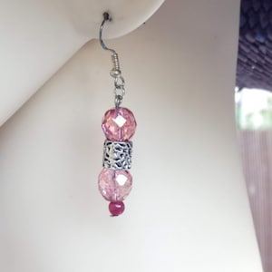 Può includere: Un singolo orecchino pendente con perline rosa sfaccettate e un accento in filigrana argentata. L'orecchino è sospeso a un gancio e una catena d'argento. Una piccola perla rossa è in basso. Sfondo neutro.