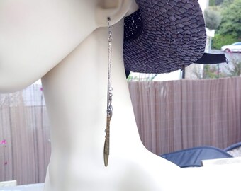 May include: A pair of long silver chain earrings with a gold leaf pendant. The earrings are hanging from a mannequin's ear.