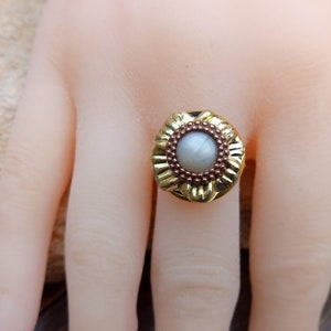 May include: A gold-colored ring with a central, light-colored stone. The ring has a floral design around the stone and a dark band. The ring is on a mannequin hand.