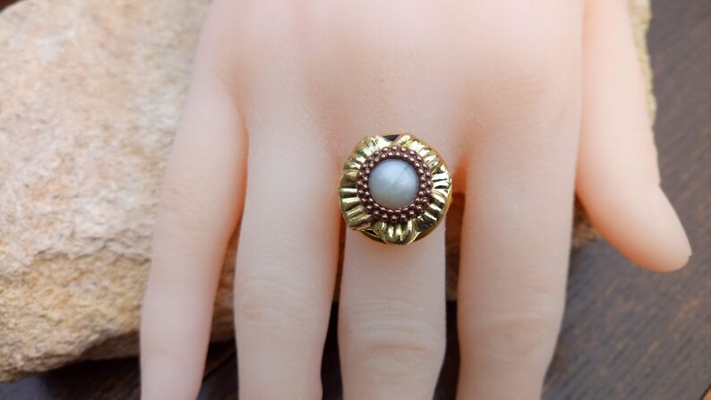 May include: A gold-colored ring with a central, light-colored stone. The ring has a floral design around the stone and a dark band. The ring is on a mannequin hand.