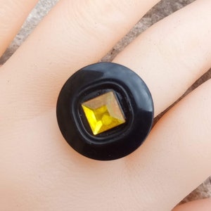 Puede incluir: Un anillo negro con una gema amarilla cuadrada engastada en un dise&ntilde;o circular. El anillo es una pieza audaz, con un acabado brillante y un contraste llamativo entre la banda negra y la vibrante gema.