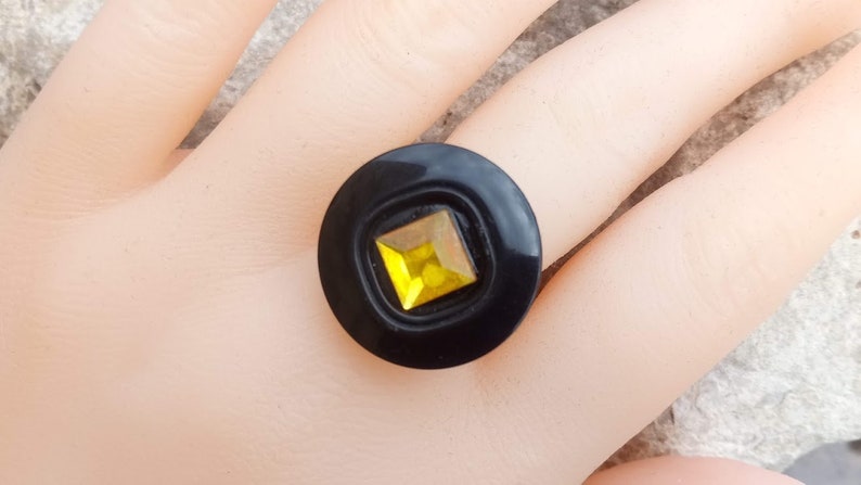 Puede incluir: Un anillo negro con una gema amarilla cuadrada engastada en un dise&ntilde;o circular. El anillo es una pieza audaz, con un acabado brillante y un contraste llamativo entre la banda negra y la vibrante gema.