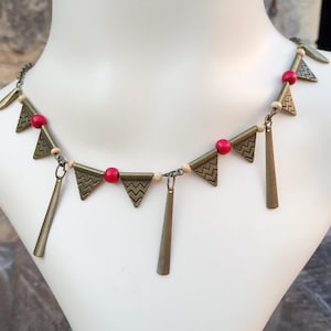 Puede incluir: Un collar de oro con cuentas rojas y blancas y colgantes triangulares. El collar está en un maniquí blanco.