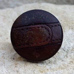 Op de afbeelding: Een ronde, bruine leren knoop met een gestikt ontwerp. De knoop heeft een donkerbruin midden en een lichtbruine buitenring.