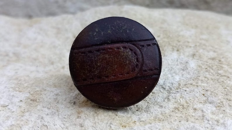 Op de afbeelding: Een ronde, bruine leren knoop met een gestikt ontwerp. De knoop heeft een donkerbruin midden en een lichtbruine buitenring.