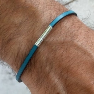 May include: A teal leather bracelet with a silver clasp is shown on a wrist. The bracelet is a thin band of teal leather with a silver magnetic clasp. The bracelet is a simple, minimalist design.