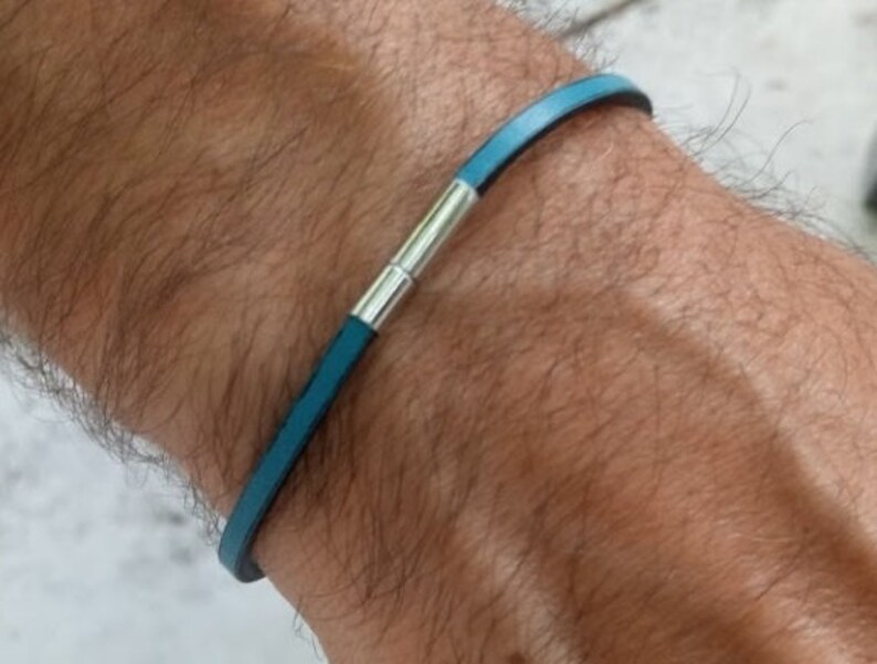 May include: A teal leather bracelet with a silver clasp is shown on a wrist. The bracelet is a thin band of teal leather with a silver magnetic clasp. The bracelet is a simple, minimalist design.