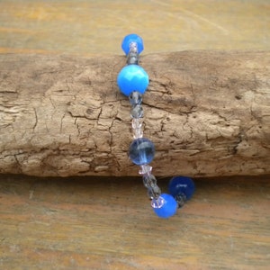Puede incluir: Un brazalete de cuentas de vidrio azul y transparente con un cierre plateado. El brazalete est&aacute; sobre un trozo de madera flotante.