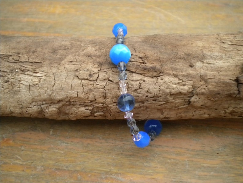 Puede incluir: Un brazalete de cuentas de vidrio azul y transparente con un cierre plateado. El brazalete est&aacute; sobre un trozo de madera flotante.
