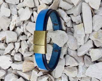 May include: A blue leather bracelet with a gold magnetic clasp. The bracelet is photographed on a bed of white gravel.