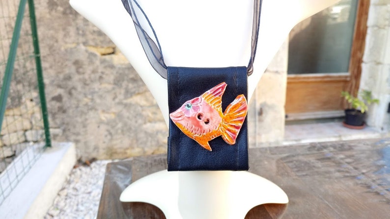Pu&ograve; includere: Una collana in pelle nera con un ciondolo in ceramica a forma di pesce arancione e rosa. Il pesce ha un bottone per l'occhio.