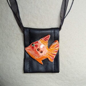Pu&ograve; includere: Una collana di pelle nera con un piccolo ciondolo a forma di pesce arancione e rosa. Il pesce ha un occhio verde ed &egrave; fatto di ceramica.