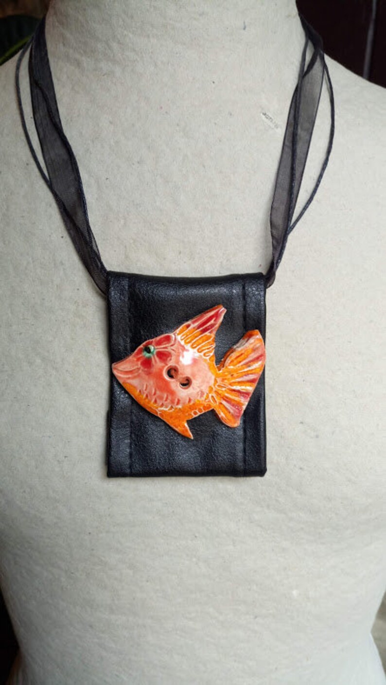 Pu&ograve; includere: Una collana di pelle nera con un piccolo ciondolo a forma di pesce arancione e rosa. Il pesce ha un occhio verde ed &egrave; fatto di ceramica.