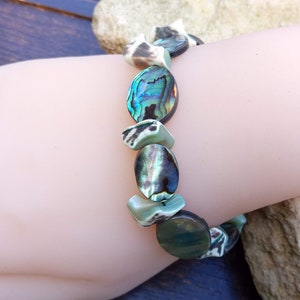 Op de afbeelding: Een armband gemaakt van iriserende groene en bruine abalone schelp kralen. De armband zit om een pols.