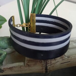 May include: A navy blue leather bracelet with two white stripes. The bracelet has a gold clasp.