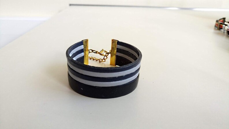 May include: A black leather bracelet with two white stripes and a gold chain closure.