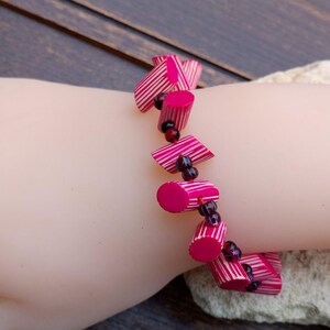 May include: A handmade bracelet featuring cylindrical and angled beads in shades of pink and white, interspersed with small, dark red beads. The bracelet is displayed on a mannequin hand.