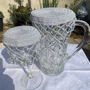 May include: Two crocheted covers for a glass jug and a wine glass. The covers are white and have a beaded fringe. The jug and glass are clear.