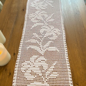 May include: White crocheted table runner with a floral design. The runner is made of delicate, openwork stitches and features a repeating pattern of flowers.