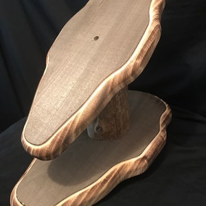 May include: A two-tiered wooden display stand with a natural edge. The top and bottom platforms are gray, with a light brown, wavy edge. The stand is supported by a natural wood post. The background is black.
