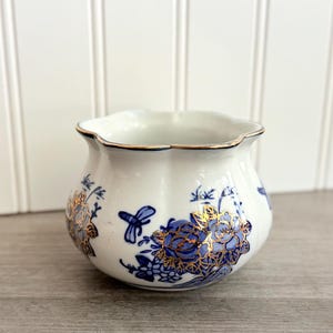 May include: A white ceramic planter with a scalloped rim and gold trim. The planter features blue floral and gold leaf designs. A blue butterfly is also visible on the planter's surface. The planter is sitting on a wooden surface.