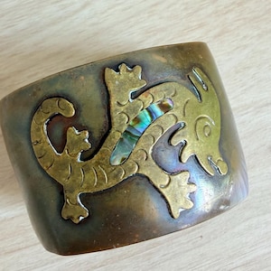 Op de afbeelding: Een brede koperen armband met een uitgesneden hagedis- of gekkomotief. Het midden van het hagedissenlichaam is ingelegd met een strook iriserende abalone-schelp. Het koper heeft een donkere, verouderde patina met zichtbare etsdetails.