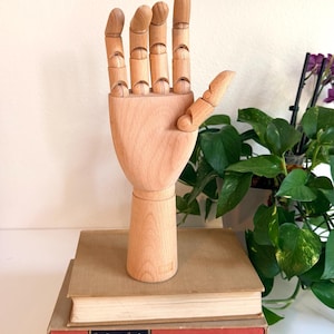 May include: A wooden articulated hand model, posed with the thumb and index finger extended. The hand is light brown and sits atop a stack of vintage books with tan, red, and gray covers. The background includes a framed print and greenery.