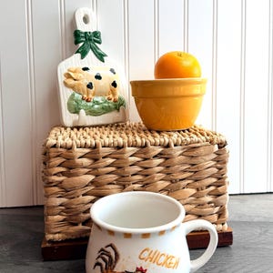 Op de afbeelding: Een stilleven met een witte keramische mok met de tekst "Chicken Soup" en een illustratie van een haan. Een gele kom bevat een sinaasappel en een varkensvormige snijplank met een groene strik is ook aanwezig. De items rusten op een geweven mand.