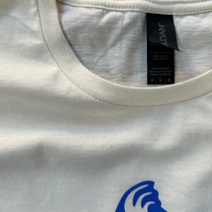 K&ouml;nnte beinhalten: Ein wei&szlig;es T-Shirt mit einem blauen Wellendesign und einem schwarzen Etikett mit der Aufschrift "ALDAN" mit dem Text "100% Baumwolle / coton / algod&oacute;n" und "Hergestellt in Bangladesch / Fabriqu&eacute; au Bangladesh / Hecho en Bangladesh" sowie den Gr&ouml;&szlig;enoptionen M, L, XL und XXL.