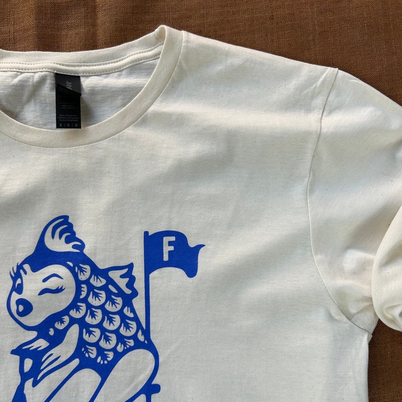 K&ouml;nnte beinhalten: Wei&szlig;es Langarm-T-Shirt mit einer blauen Grafik eines Cartoon-Fisches, der eine blaue Flagge mit dem Buchstaben "F" h&auml;lt.