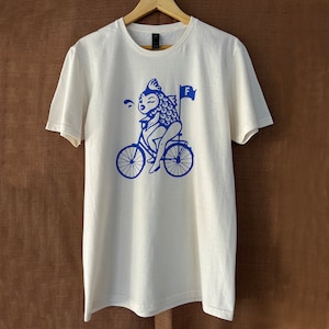 K&ouml;nnte beinhalten: Wei&szlig;es T-Shirt mit einem blauen Motiv eines Fisches, der ein Fahrrad f&auml;hrt, mit dem Buchstaben "F" auf einer Flagge.
