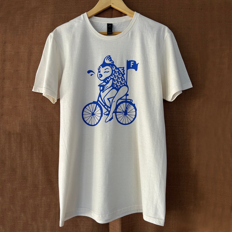 K&ouml;nnte beinhalten: Wei&szlig;es T-Shirt mit einem blauen Motiv eines Fisches, der ein Fahrrad f&auml;hrt, mit dem Buchstaben "F" auf einer Flagge.