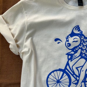 K&ouml;nnte beinhalten: Ein wei&szlig;es T-Shirt mit einem blauen Motiv eines Cartoon-Fischs, der Fahrrad f&auml;hrt.