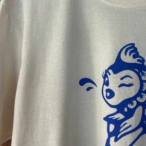 K&ouml;nnte beinhalten: Wei&szlig;es T-Shirt mit einem blauen Grafikdruck eines Comic-Charakters mit einem zwinkernden Auge und einer einzigen Haarlocke. Der Charakter bl&auml;st eine Blase.