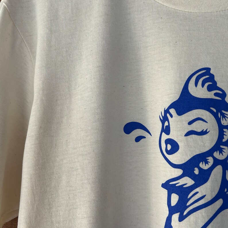 K&ouml;nnte beinhalten: Wei&szlig;es T-Shirt mit einem blauen Grafikdruck eines Comic-Charakters mit einem zwinkernden Auge und einer einzigen Haarlocke. Der Charakter bl&auml;st eine Blase.