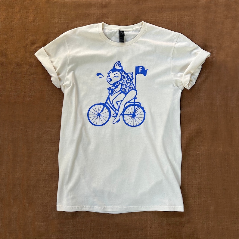 Feminist T-shirt with illustration depicting the iconic Irina Dunn feminism quote "A woman needs a man like a fish needs a bicycle." Printed in the USA with blue / periwinkle ink on a natural fiber shirt. Woman owned business.