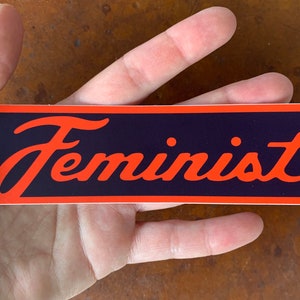 以下が含まれることがあります： 赤い縁取りのある黒い背景にオレンジ色の「フェミニスト」という文字が書かれた長方形のステッカー。ステッカーは光沢仕上げです。