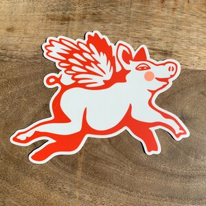 May include: A red and white illustration of a pig with wings flying. The pig has a curled tail and a pink blush on its cheek.