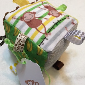 May include: A soft, multi-textured baby toy cube with a monkey theme. The cube has different fabrics and textures, including a green fabric with bananas, a white fabric with striped monkeys, and a gray fabric. The cube has a brown tag with a monkey illustration and the text "Monkey".