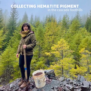 Puede incluir: Una persona con una chaqueta verde y una bufanda est&aacute; de pie en un bosque sosteniendo una pala. Un cubo lleno de tierra roja se encuentra en el suelo frente a ella. El texto "COLLECTING HEMATITE PIGMENT in the cascade foothills" est&aacute; en la parte superior de la imagen.