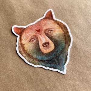 Puede incluir: Una ilustración de acuarela de la cara de un oso con un pequeño corazón rojo en la frente. El oso tiene un pelaje marrón con matices azules y verdes. La ilustración está rodeada por un borde blanco con el texto "WHITESNAKE ARTS" en blanco.