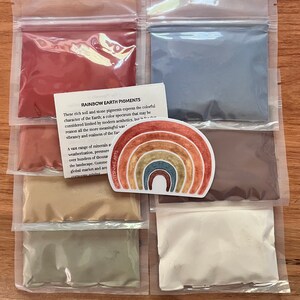 Puede incluir: Seis peque&ntilde;as bolsas de pl&aacute;stico llenas de pigmentos de tierra de diferentes colores. Las bolsas est&aacute;n dispuestas en dos filas de tres. Los pigmentos son rojo, azul, marr&oacute;n, beige, verde y blanco. Una pegatina de arco&iacute;ris con el texto "WHITESNAKE ARTS" est&aacute; en el centro de la imagen. El texto "RAINBOW EARTH PIGMENTS" est&aacute; en una tarjeta blanca en la esquina superior izquierda de la imagen. El texto dice: "Estos ricos pigmentos de tierra y piedra expresan el car&aacute;cter colorido de la Tierra; un espectro de colores que puede considerarse limitado por la est&eacute;tica moderna, pero por esa raz&oacute;n, a&uacute;n m&aacute;s significativo para expresar la vitalidad y la realidad de la Tierra."
