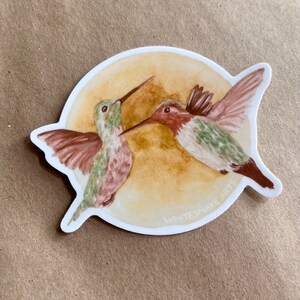 Puede incluir: Una pegatina con una ilustración de acuarela de dos colibríes en vuelo sobre un fondo amarillo. La pegatina tiene el texto "WHITESNAKE ARTS" en la parte inferior.
