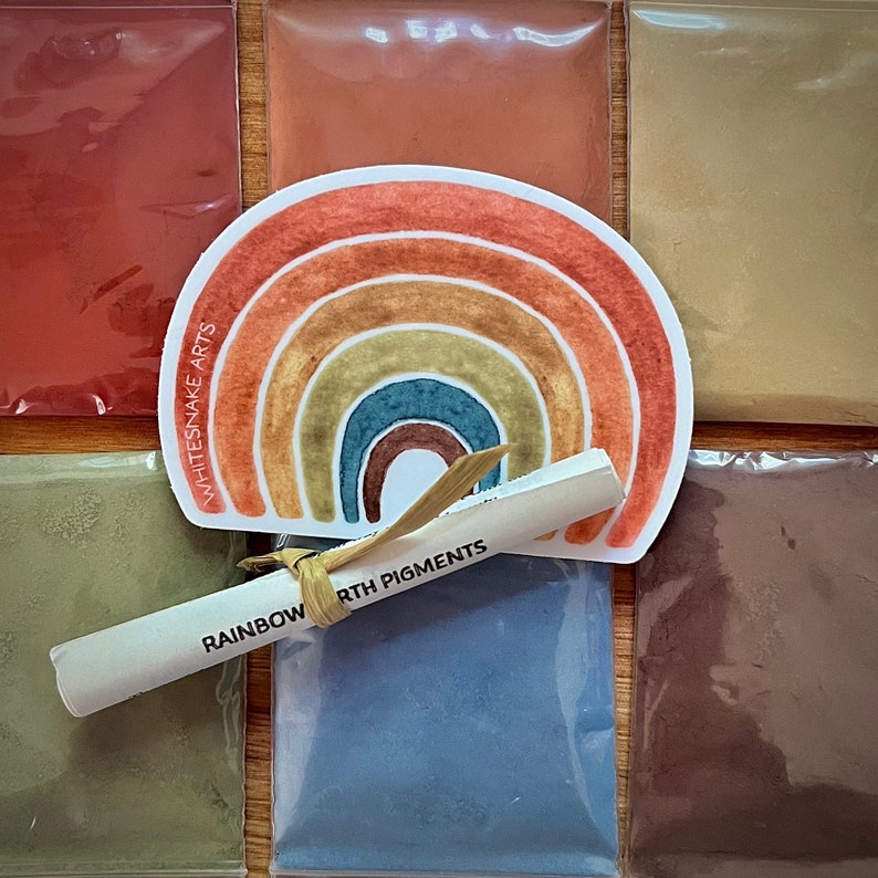 Puede incluir: Un conjunto de siete peque&ntilde;as bolsas de pigmentos de tierra, cada una de un color diferente. Las bolsas est&aacute;n dispuestas en fila, con una ilustraci&oacute;n de arco&iacute;ris y el texto "Rainbow Earth Pigments" en una etiqueta blanca delante de ellas.