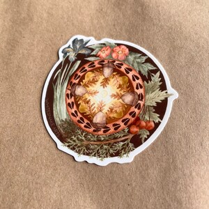 Puede incluir: Una pegatina redonda con fondo marrón que presenta un diseño tipo mandala con bellotas, hojas y flores. El texto "WHITESNAKE ARTS" está escrito verticalmente en el lado izquierdo de la pegatina.