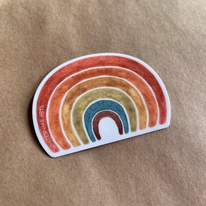 Puede incluir: Una pegatina con un arcoíris de acuarela en tonos rojos, naranjas, amarillos, verdes, azules y marrones. La pegatina tiene el texto "WHITESNAKE ARTS" en el lado izquierdo.